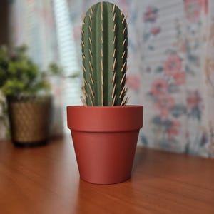 May include: A decorative cactus in a terracotta-colored pot. The cactus is a vibrant green with light brown spines. The pot is a classic, simple design, adding a touch of desert charm to any space. The background is blurred, with a floral curtain.