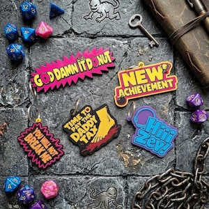 Może przedstawiać: Pięć kolorowych breloczków z napisami, w tym GOD DAMN IT DONUT, NEW ACHIEVEMENT, YOU WILL NOT BREAK ME, TIME TO PAY THE DADDY TAX oraz Hiii Zev!, w otoczeniu różnych wielościennych kości do gry, metalowego klucza i metalowego łańcucha.