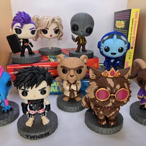 May include: A collection of eight stylised vinyl figures featuring diverse characters, including a blue dinosaur, a punk-inspired figure, a blonde character, a grey alien, a lion-like creature, a blue robot, a cat with glasses, and a goat-like figure.