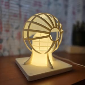May include: A cream-colored globe-shaped lamp with a grid design, emitting a warm glow. The lamp sits on a square base and has a cord for power. The lamp is a decorative lighting fixture.