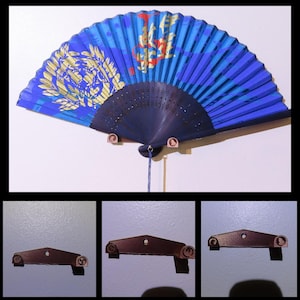 May include: A blue and black hand fan with a decorative design, mounted on a white wall. The fan features a gold emblem and red accents. The fan is held in place by three brown brackets.