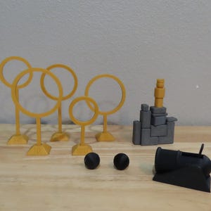 May include: A collection of 3D-printed toys, including yellow Quidditch hoops, a gray castle, two black balls, and a black cannon. The toys are arranged on a wooden surface.