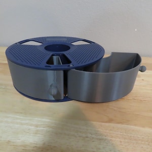 May include: A 3D-printed filament spool holder in gray and blue. The holder has a perforated blue lid and a separate gray compartment. The design includes a side compartment and a spool-holding section.