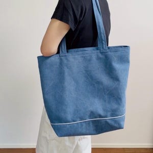 Op de afbeelding: Een blauwe tote bag met bijpassende handvatten en een witte accentlijn aan de onderkant. De tas is gemaakt van een textuurrijk materiaal en wordt over de schouder gedragen.