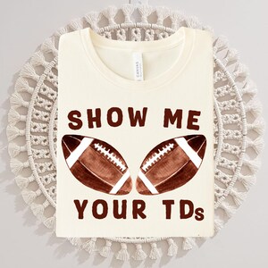 以下が含まれることがあります： クリーム色のTシャツ。茶色で「SHOW ME YOUR TDs」の文字と、2つの茶色のフットボールのイラストが描かれています。柔らかい素材でできており、カジュアルな服装に適しています。