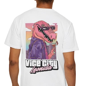 Pode incluir: Uma t-shirt branca com um gráfico nas costas de um jacaré cor-de-rosa a usar óculos de sol e uma camisa roxa estampada, segurando um cocktail. O fundo mostra o horizonte de uma cidade ao pôr do sol. O texto diz Vice City Leonida em letras.