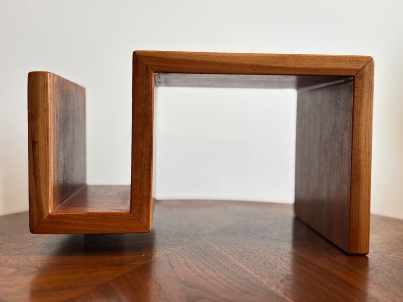 Puede incluir: Una estanter&iacute;a de madera con un dise&ntilde;o abierto &uacute;nico. La estanter&iacute;a est&aacute; construida con madera de tonos c&aacute;lidos, con un marco rectangular y un soporte vertical. El dise&ntilde;o es moderno y minimalista, adecuado para exhibir libros o decoraci&oacute;n.