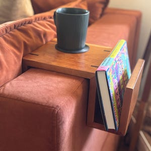 Puede incluir: Una mesa de brazo de sof&aacute; de madera con un sujetalibros incorporado y una taza negra. La mesa est&aacute; colocada sobre un sof&aacute; de color &oacute;xido. Un libro colorido est&aacute; en el soporte.