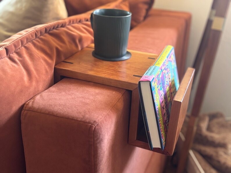 Puede incluir: Una mesa de brazo de sof&aacute; de madera con un sujetalibros incorporado y una taza negra. La mesa est&aacute; colocada sobre un sof&aacute; de color &oacute;xido. Un libro colorido est&aacute; en el soporte.