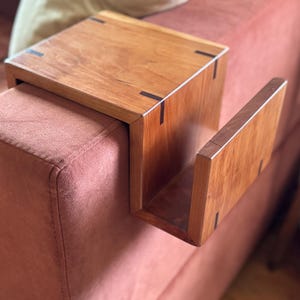 Puede incluir: Una mesa de brazo de sof&aacute; de madera con un revistero incorporado. La mesa es de madera marr&oacute;n claro con detalles en marr&oacute;n oscuro. Se coloca sobre un sof&aacute; rosa. La mesa est&aacute; dise&ntilde;ada para encajar sobre el brazo de un sof&aacute;.