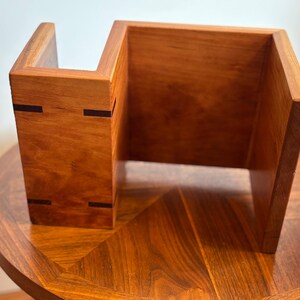 Puede incluir: Un organizador de escritorio de madera hecho a mano con un c&aacute;lido acabado marr&oacute;n rojizo. El organizador presenta un dise&ntilde;o &uacute;nico y abierto con una forma escalonada y detalles oscuros. Se encuentra sobre una mesa redonda de madera con una superficie estampada.