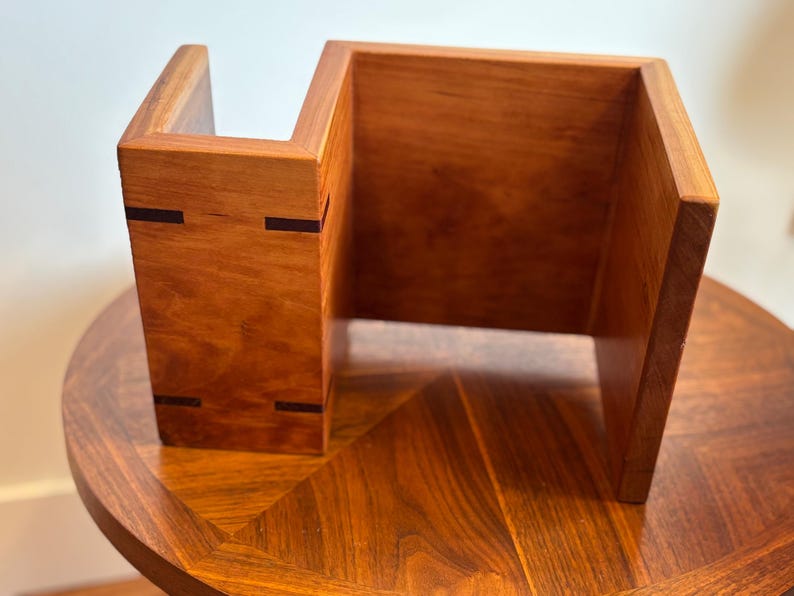Puede incluir: Un organizador de escritorio de madera hecho a mano con un c&aacute;lido acabado marr&oacute;n rojizo. El organizador presenta un dise&ntilde;o &uacute;nico y abierto con una forma escalonada y detalles oscuros. Se encuentra sobre una mesa redonda de madera con una superficie estampada.