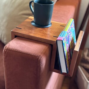 Puede incluir: Una mesa de brazo de sof&aacute; de madera con un sujetalibros incorporado y una taza de color verde oscuro. La mesa es de madera de tonos c&aacute;lidos y est&aacute; sujeta a un sof&aacute; de color &oacute;xido. Un libro colorido est&aacute; en el soporte.