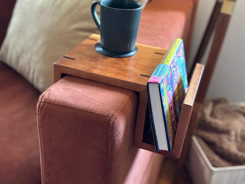 Puede incluir: Una mesa de brazo de sof&aacute; de madera con un sujetalibros incorporado y una taza de color verde oscuro. La mesa es de madera de tonos c&aacute;lidos y est&aacute; sujeta a un sof&aacute; de color &oacute;xido. Un libro colorido est&aacute; en el soporte.