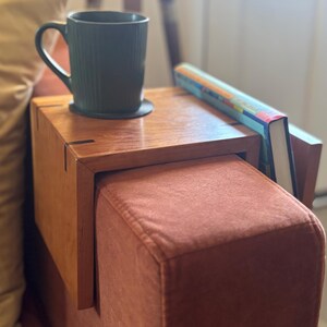 Puede incluir: Una mesa de apoyabrazos de madera con una taza verde oscuro y un libro. La mesa est&aacute; sujeta a un reposabrazos tapizado de color &oacute;xido. La taza est&aacute; sobre un posavasos negro. El libro tiene una cubierta colorida.