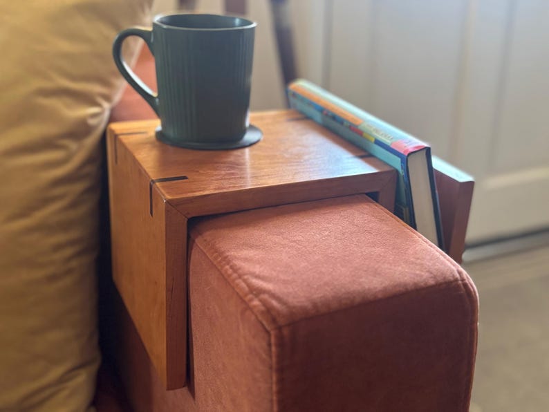 Puede incluir: Una mesa de apoyabrazos de madera con una taza verde oscuro y un libro. La mesa est&aacute; sujeta a un reposabrazos tapizado de color &oacute;xido. La taza est&aacute; sobre un posavasos negro. El libro tiene una cubierta colorida.