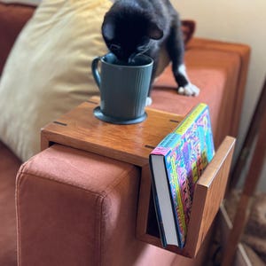 Puede incluir: Una mesa de brazo de sof&aacute; de madera con un sujetalibros incorporado. Una taza verde azulado est&aacute; sobre la mesa, y un libro con una cubierta colorida est&aacute; en el soporte. Un gato negro est&aacute; cerca de la taza. El sof&aacute; es de color &oacute;xido.