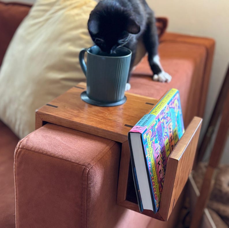 Puede incluir: Una mesa de brazo de sof&aacute; de madera con un sujetalibros incorporado. Una taza verde azulado est&aacute; sobre la mesa, y un libro con una cubierta colorida est&aacute; en el soporte. Un gato negro est&aacute; cerca de la taza. El sof&aacute; es de color &oacute;xido.