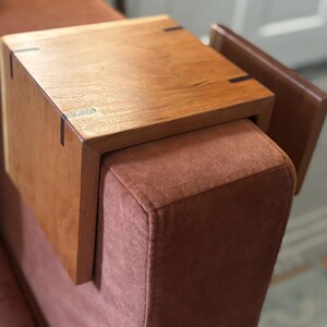 Puede incluir: Una mesa de apoyabrazos de madera en un tono marr&oacute;n c&aacute;lido, dise&ntilde;ada para encajar sobre un brazo de sof&aacute;. La mesa presenta una parte superior rectangular con vetas de madera visibles y uniones de cola de milano. El brazo del sof&aacute; est&aacute; tapizado en tela marr&oacute;n rojizo.