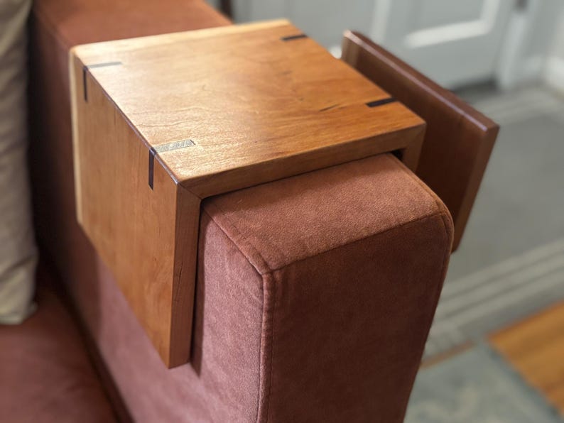 Puede incluir: Una mesa de apoyabrazos de madera en un tono marr&oacute;n c&aacute;lido, dise&ntilde;ada para encajar sobre un brazo de sof&aacute;. La mesa presenta una parte superior rectangular con vetas de madera visibles y uniones de cola de milano. El brazo del sof&aacute; est&aacute; tapizado en tela marr&oacute;n rojizo.