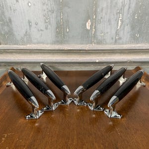 May include: A set of six cabinet handles featuring a sleek, elongated black centre section with polished chrome-finished metal ends and mounting brackets. Each handle is designed with a curved, ergonomic shape and includes two screw holes for attachment.