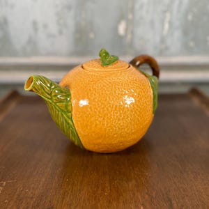 May include: A ceramic teapot shaped like a textured orange fruit. The body features a bright orange, dimpled surface with a green leaf accent on the spout and lid. A brown curved handle is attached to the side, completing the fruit-inspired design.