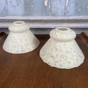 May include: A pair of bell-shaped glass lamp shades featuring a creamy off-white base colour with a mottled, amber-brown speckled pattern throughout. Each shade has a rounded top collar and a wider, flared bottom opening with a smooth edge.
