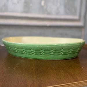 May include: Oval ceramic baking dish with a light yellow interior and a green exterior. The rim has a wave pattern. The dish sits on a wooden surface.