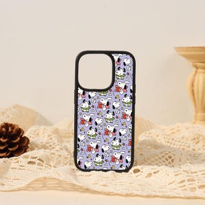 May include: A black phone case featuring a repeating pattern of the cartoon character Snoopy on a light purple background. The design includes multiple illustrations of the white dog holding a red heart, surrounded by small red stars and squiggly lines.