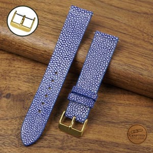 May include: A blue watch strap with a textured, pebbled surface pattern and a gold-toned metal buckle. The strap consists of two pieces, one with a series of adjustment holes and the other featuring a matching loop and a gold-toned pin buckle.
