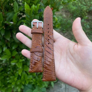 Puede incluir: Una correa de reloj de cuero marrón con una superficie texturizada y costuras en contraste. La correa presenta una hebilla plateada y está diseñada para un reloj. El cuero tiene un patrón único.