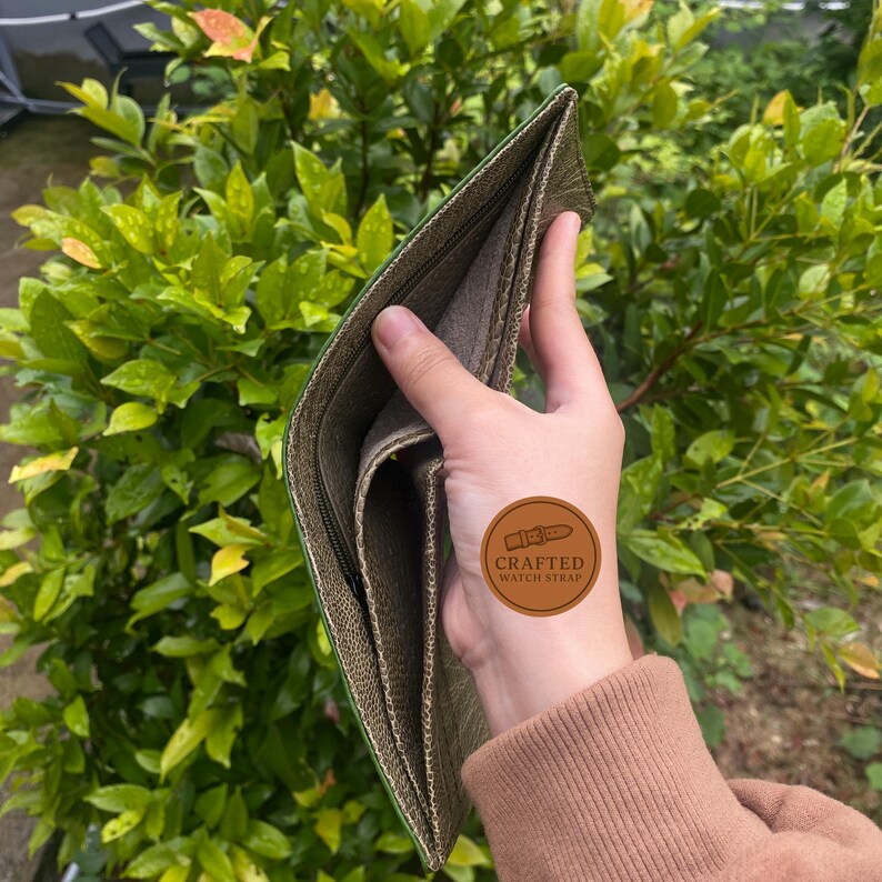 Puede incluir: Cartera verde oliva abierta con cierre de cremallera, sobre un fondo de follaje verde. El interior de la cartera es de un verde m&aacute;s claro. Una pegatina marr&oacute;n con el texto "CRAFTED WATCH STRAP" es visible.