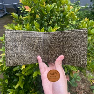 Puede incluir: Cartera de cuero verde oliva abierta, con m&uacute;ltiples ranuras para tarjetas. La cartera tiene una superficie texturizada y se sostiene abierta, mostrando su interior. El logotipo "Crafted Watch Strap" es visible.