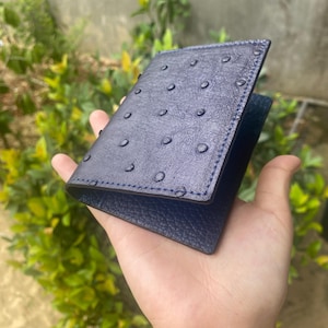 Puede incluir: Una cartera de cuero azul marino con una superficie texturizada y un borde cosido. La cartera está abierta, revelando un interior azul oscuro. La cartera se sostiene en una mano.