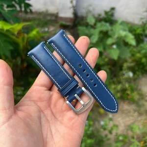 Pode incluir: Um par de pulseiras de relógio em couro azul marinho com costura branca. As pulseiras têm uma fivela de aço inoxidável e são seguradas em uma mão contra um fundo de folhagem verde.