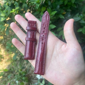 Puede incluir: Un par de correas de reloj burdeos con un patrón texturizado similar al de la piel de cocodrilo. Una correa tiene una hebilla y la otra tiene varios agujeros para ajustar. Las correas están hechas de un material flexible.