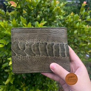 Puede incluir: Una cartera de cuero verde con una superficie texturizada y un patr&oacute;n distintivo. La cartera se sostiene en una mano, mostrando su dise&ntilde;o y artesan&iacute;a. El color de la cartera es un verde apagado, y la textura a&ntilde;ade inter&eacute;s visual.