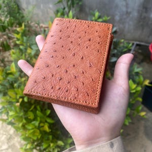 Peut inclure: Un portefeuille en cuir marron avec une surface texturée, tenu dans une main. Le portefeuille est de forme rectangulaire avec des coins arrondis et des coutures visibles. Le cuir présente un motif distinctif de petites bosses en relief.