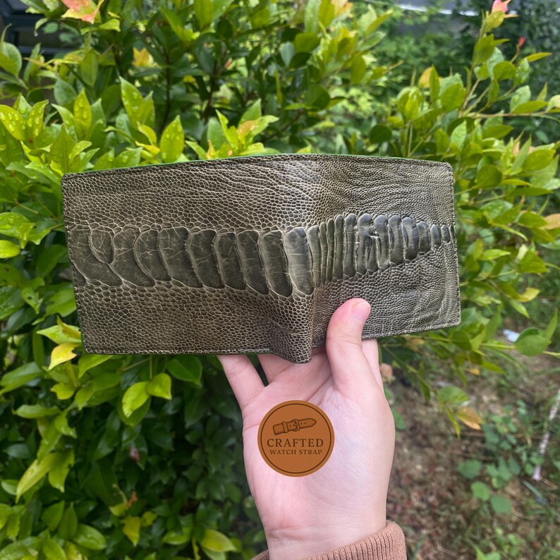 Puede incluir: Cartera de cuero verde oliva con dise&ntilde;o de pata de avestruz texturizada. La cartera se sostiene en una mano, mostrando el patr&oacute;n y color &uacute;nicos. La cartera tiene un dise&ntilde;o de dos pliegues y parece nueva.
