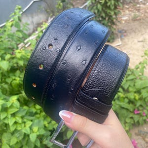 Puede incluir: Un cinturón negro con una superficie texturizada que presenta un patrón de pequeñas hendiduras circulares que recuerdan al cuero de avestruz. El cinturón incluye una serie de agujeros perforados para el ajuste y una hebilla metálica.
