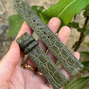 Könnte beinhalten: Olivgrünes Uhrenarmband mit Krokodilleder-Muster. Das Armband besteht aus Leder und hat weiße Nähte an den Kanten. Das Uhrenarmband wird vor einem Hintergrund aus grünen Blättern gezeigt.
