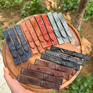 May include: A collection of watch straps in various colors including blue, orange, red, grey, black, and brown. Each strap features a textured ostrich leather pattern, a buckle, and multiple adjustment holes, arranged on a round wooden tray.