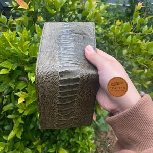 Puede incluir: Correa de reloj de cuero verde oliva con una superficie texturizada y estampada. La correa tiene un dise&ntilde;o central en relieve y una forma redondeada. El fondo es una vista borrosa de follaje verde.