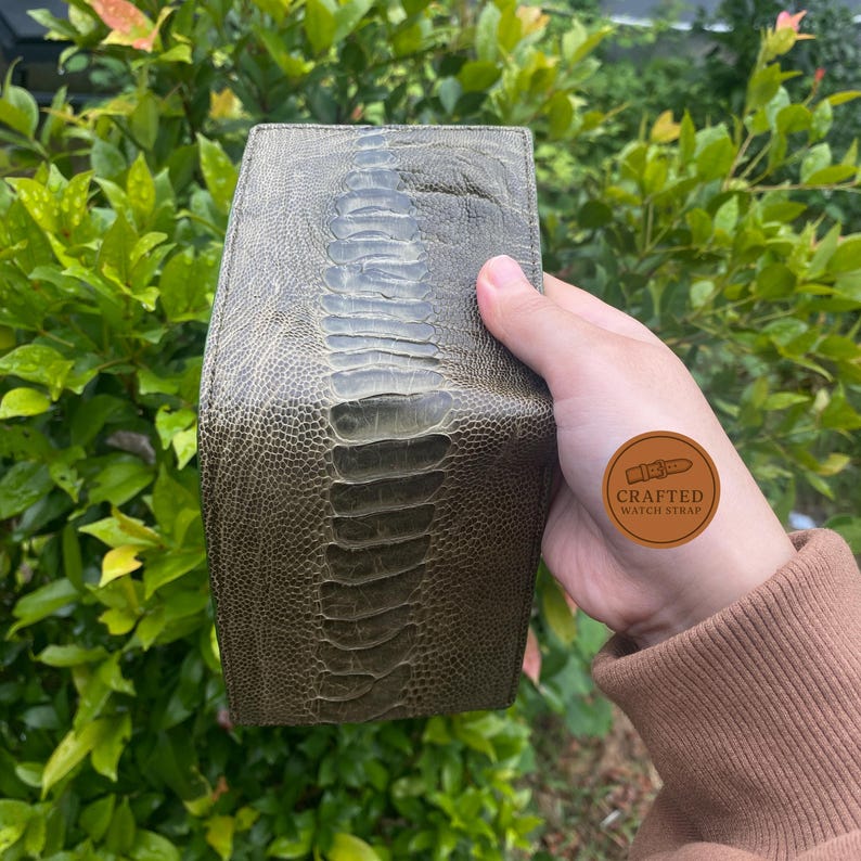 Puede incluir: Correa de reloj de cuero verde oliva con una superficie texturizada y estampada. La correa tiene un dise&ntilde;o central en relieve y una forma redondeada. El fondo es una vista borrosa de follaje verde.