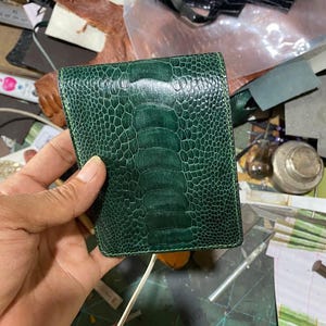 Op de afbeelding: Een donkergroene portemonnee met een textuur die op reptielenhuid lijkt. De portemonnee wordt in een hand gehouden, waarbij de kleur en het ontwerp worden benadrukt. De portemonnee lijkt van leer te zijn en is een bi-fold model.