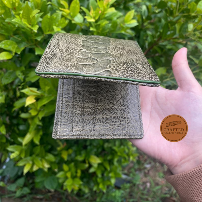 Puede incluir: Cartera de cuero de avestruz verde con superficie texturizada y ribete verde. La cartera est&aacute; abierta, revelando el interior. El logotipo de Crafted Watch Strap es visible.