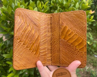Könnte beinhalten: Offene, braune Lederbrieftasche mit einem strukturierten Straußenbeinmuster. Die Brieftasche hat mehrere Kartenfächer und ein Geldscheinfach. Das Bild enthält ein rundes Logo mit der Aufschrift "Crafted Watch Strap".