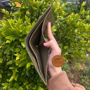 Puede incluir: Cartera de cuero verde oliva abierta con m&uacute;ltiples compartimentos. La cartera se sostiene abierta, revelando el forro interior y un bolsillo con cremallera. El cuero tiene una apariencia texturizada. Se ve un peque&ntilde;o logotipo redondo.