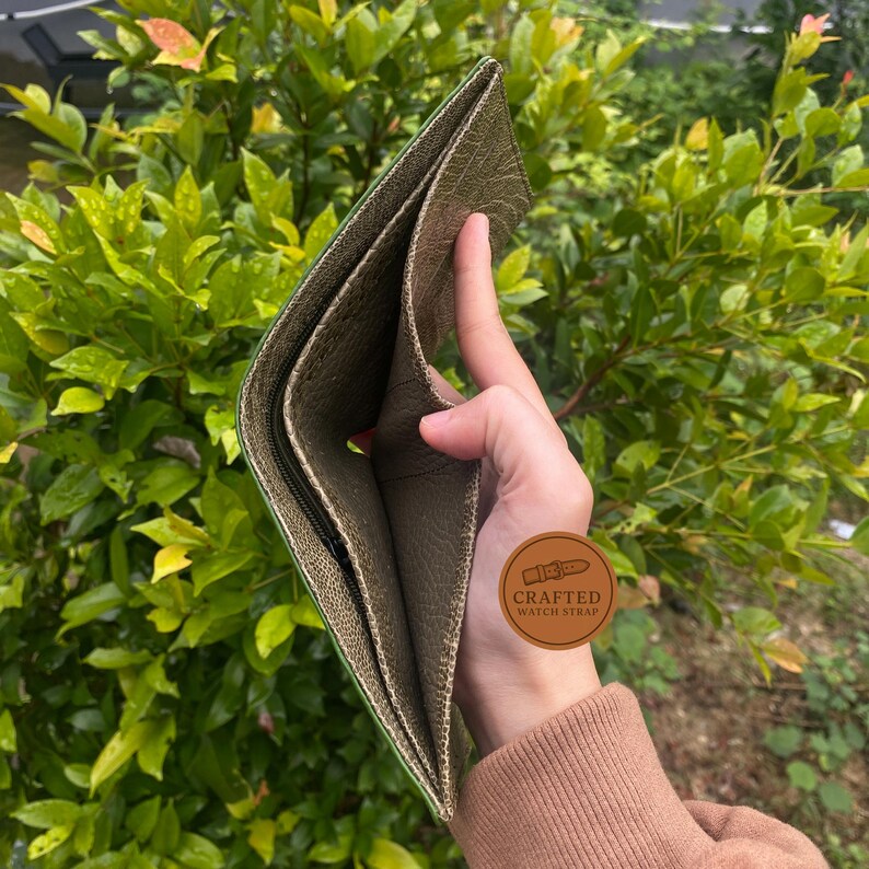 Puede incluir: Cartera de cuero verde oliva abierta con m&uacute;ltiples compartimentos. La cartera se sostiene abierta, revelando el forro interior y un bolsillo con cremallera. El cuero tiene una apariencia texturizada. Se ve un peque&ntilde;o logotipo redondo.