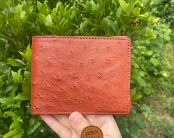 Könnte beinhalten: Ein orangefarbenes Lederportemonnaie mit einer strukturierten Straußenmusterung. Das rechteckige Portemonnaie hat einen genähten Rand. Ein kleines rundes Logo mit dem Text "Crafted Watch Strap" ist sichtbar.